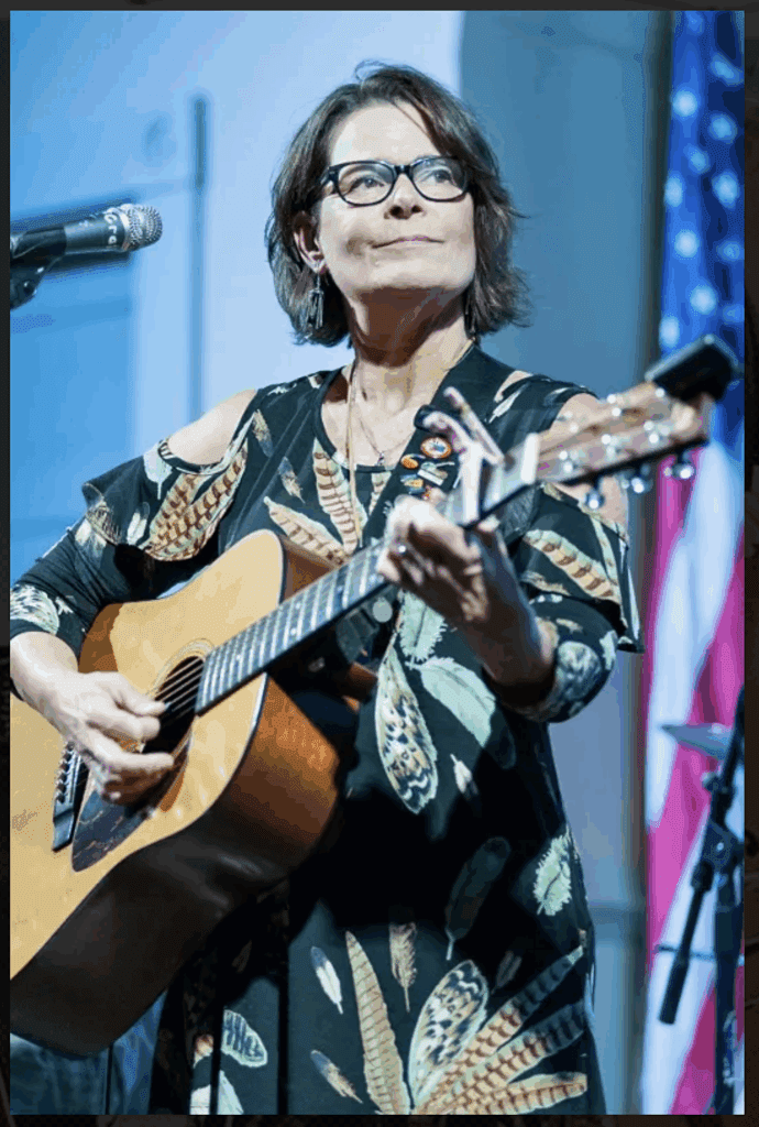 Sarah Barker Huhn performing on stage, playing an acoustic guitar and singing into a microphone, with the U.S. flag visible in the background.