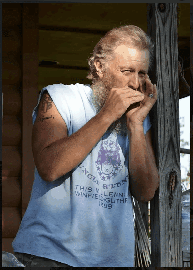 John Williams playing harmonica outdoors on a porch, wearing a light blue T-shirt and sporting a long beard.