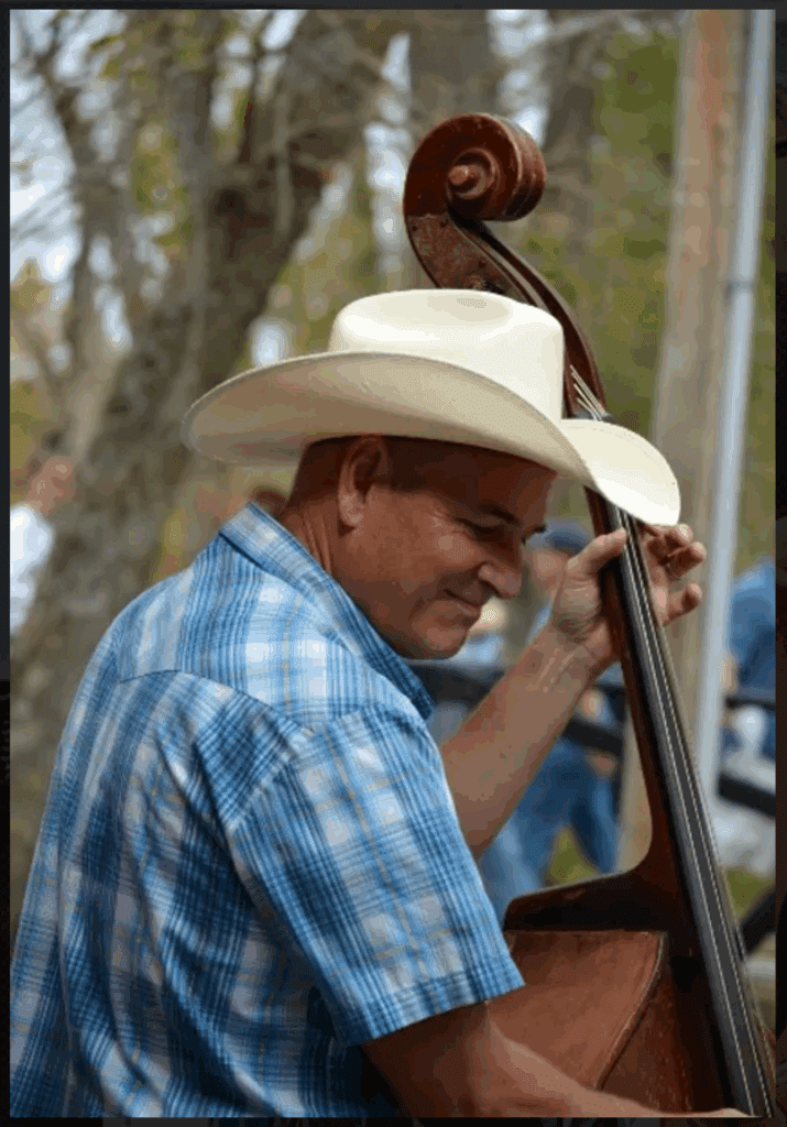 Steve Huhn playing an upright bass outdoors, wearing a cowboy hat and blue plaid shirt, focused on his performance.
