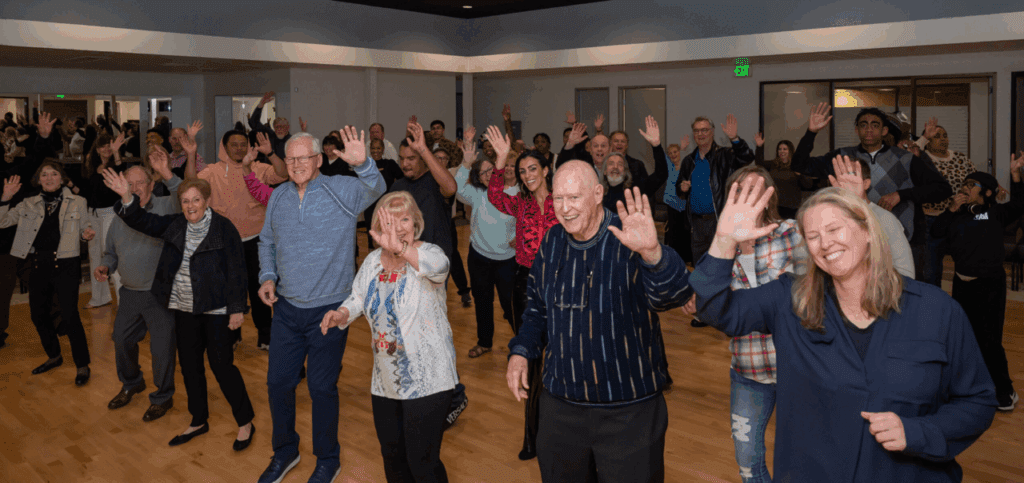 Large group of smiling people line dancing indoors, waving toward the camera, during a Classic Americana Road Show event.