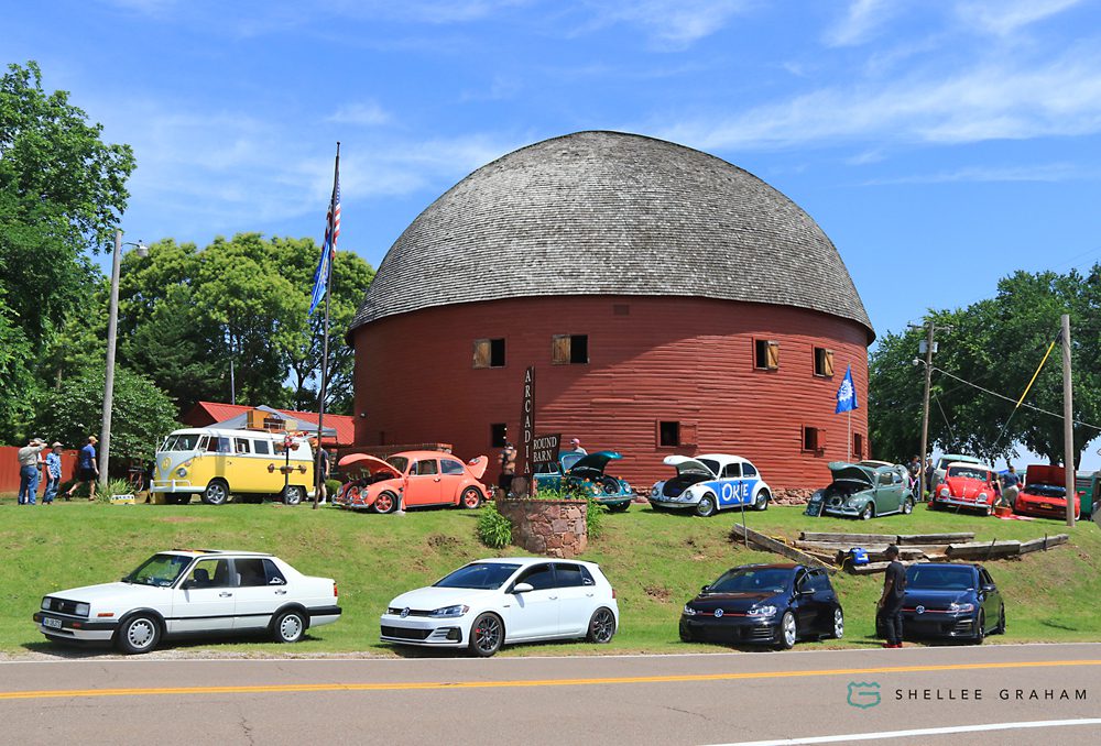 Classic cars and vintage Volkswagen Beetles displayed outside the historic Arcadia Round Barn along Route 66 in Arcadia, Oklahoma.
